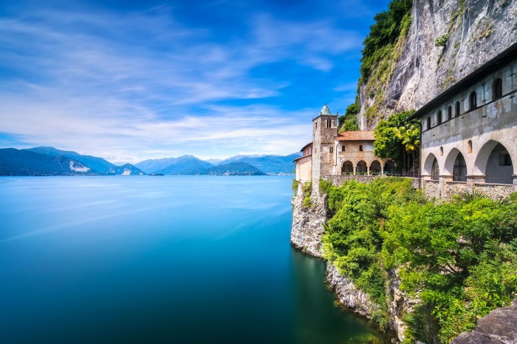 Eremo di Santa Caterina del Sasso affacciato sul Lago Maggiore, con vista panoramica su acqua e montagne in una giornata limpida.